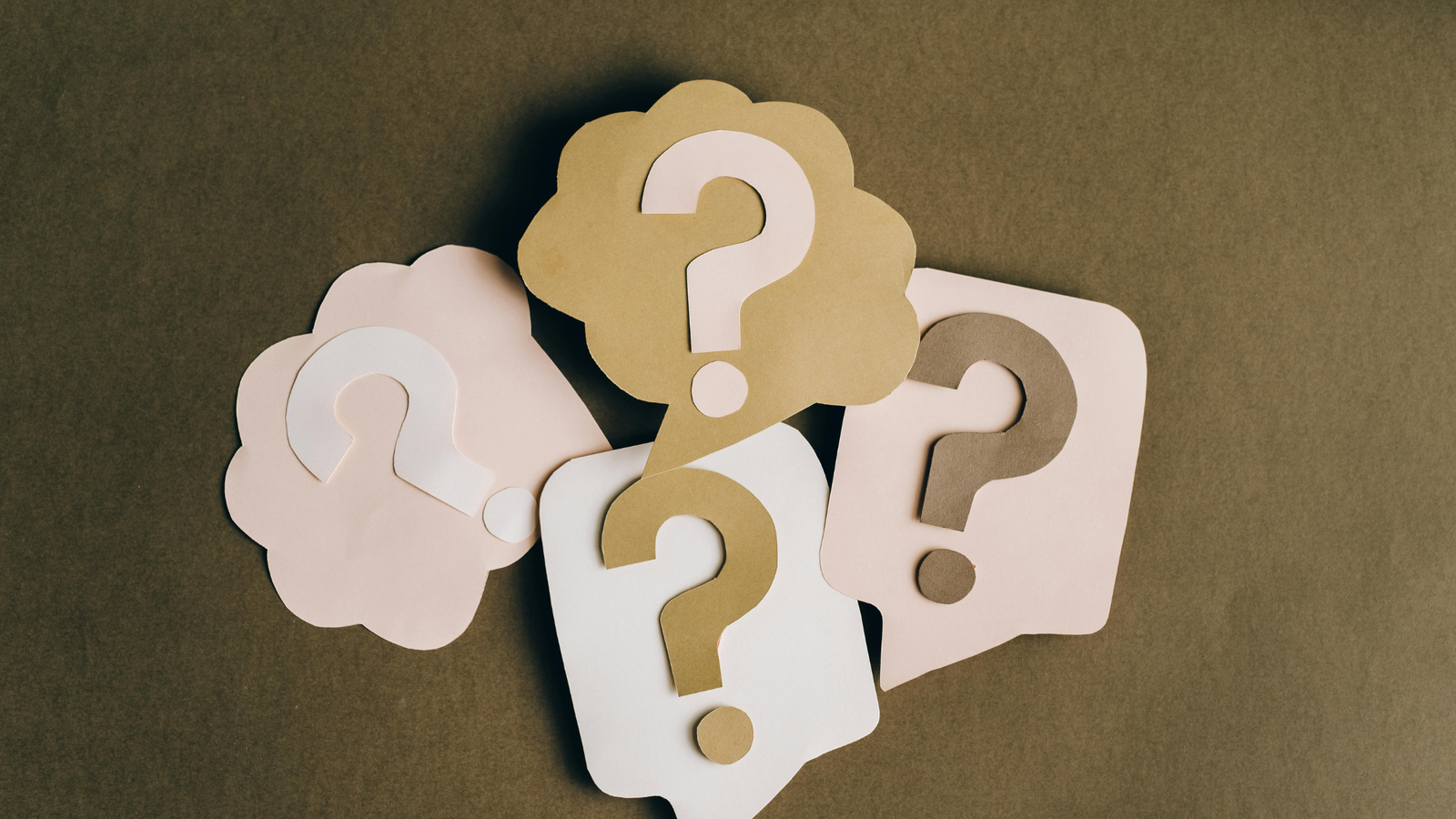 Five paper cut-out question marks in gold, white and pale pink layered on a brown background