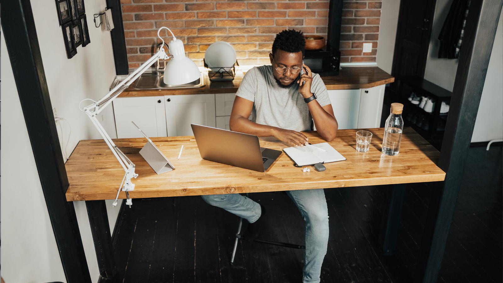 Man in glasses working from home at a wooden desk, taking a call beside a laptop and notebook in a brick walled kitchen